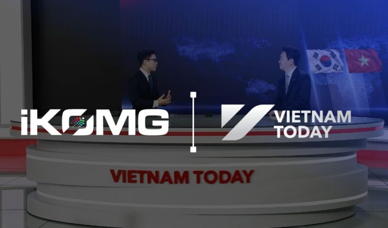 Two male news anchors sit at a circular multi-tiered desk branded 'VIETNAM TODAY' in a blue news studio with a world map backdrop and flags (South Korea and Vietnam).